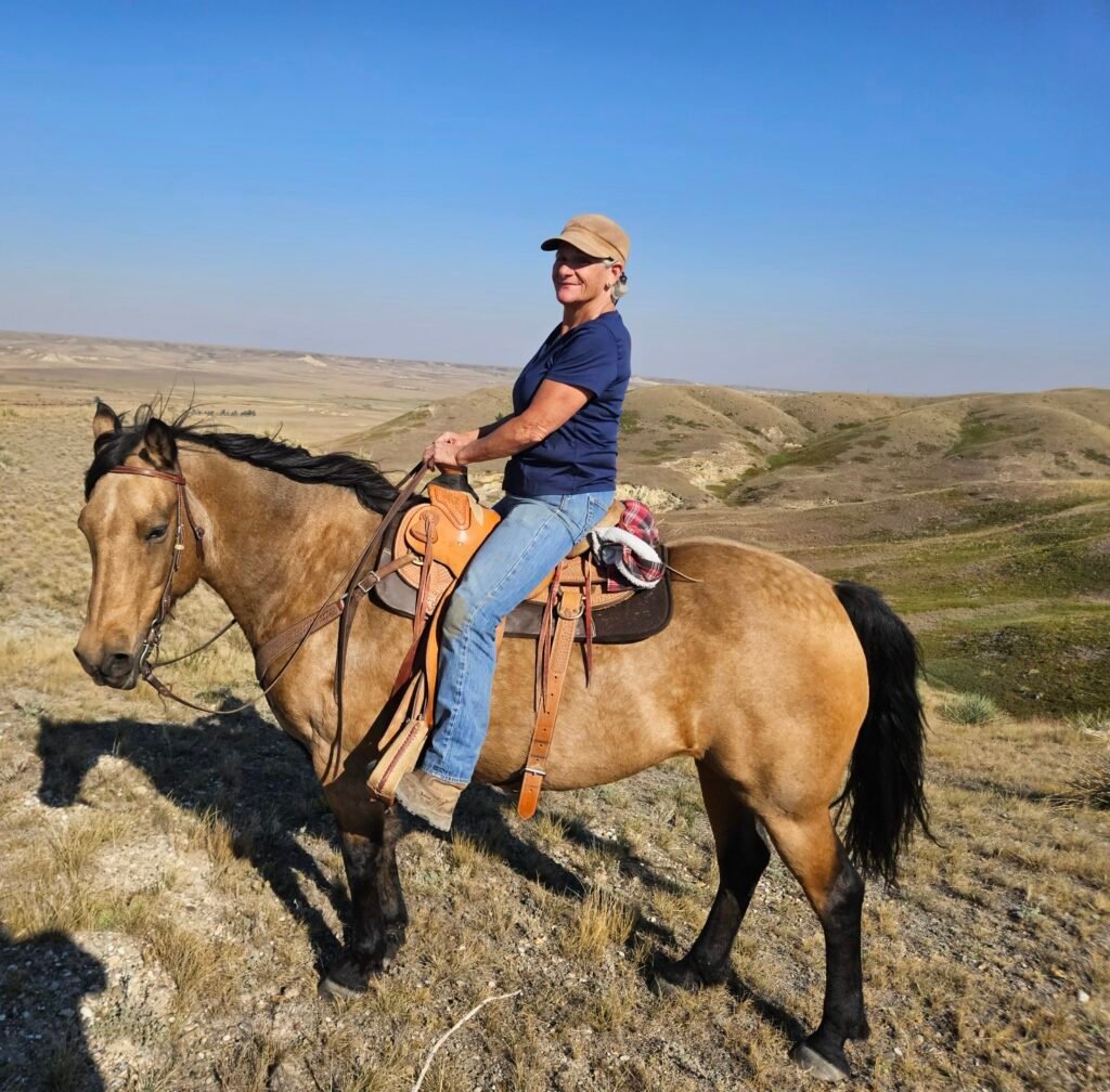 Lady in a baseball cap on a buckskin (golden-brown with black mane and tail) horse out in the country.